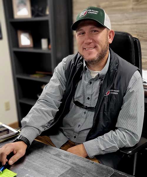 photo of Matt Simpson wearing Mid-Valley cap and vest sitting at his desk.  He has a short beard and a nice smile with a shelf in the background holding pictures.