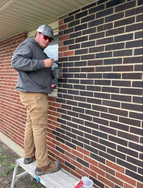 Tech staining masonry brick standing on a short platform on the ground - he is wearing a cap and gray sweatshirt with a hood - he has on sunglasses and their is a window behind him.