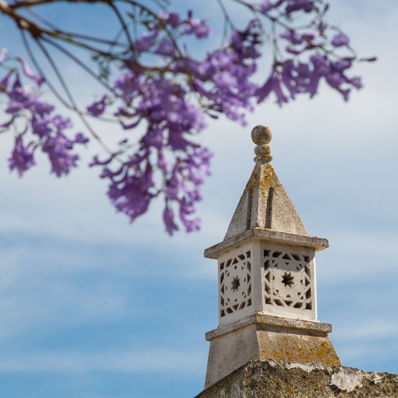 an ornate white old-fashioned chimney with purple flowers growing near it